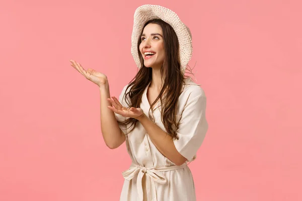 Girl enjoying sunny perfect weather on summer vacation. Cheerful attractive brunette female in dress and hat introduce something, taking look at beautiful thing, smiling, pink background — 스톡 사진