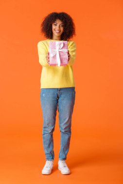 Full-length vertical shot cute friendly african-american woman, best friend stretching hands and giving lovely pink wrapped gift to you, smiling, congratulating with holidays, orange background
