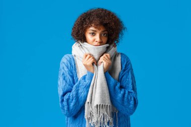Cute and gloomy young african-american lovely woman with afro haircut, wrap herself with warm scarf, feeling chilly or cold in winter, standing blue background, waiting someone fix heater