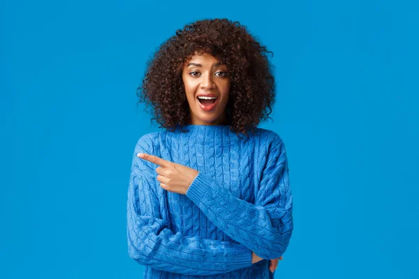 Amusée, heureuse et fascinée femme afro-américaine souriante appréciant la station de ski passer des vacances d'hiver, portant un pull bleu pointant du doigt vers la gauche et souriant excité, debout fond studio — Photo