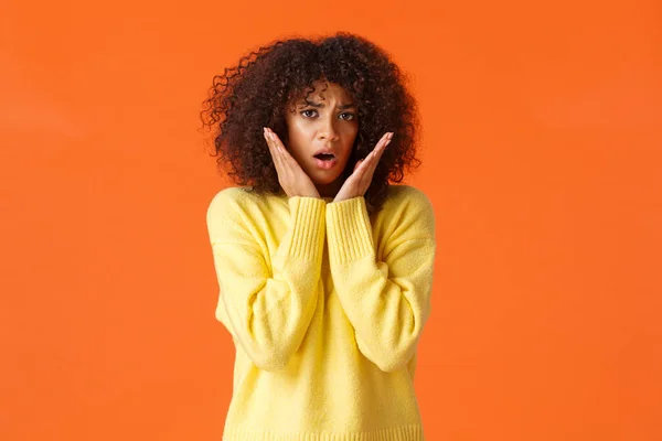 Scared timid and insecure lovely young girl with afro haircut, yellow sweater, gasping shocked and concerned, touching and staring in panic camera, feeling afraid, standing orange background — 스톡 사진