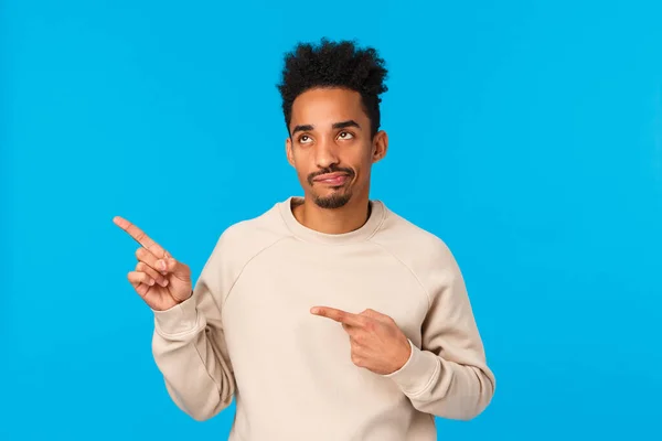 Cético e desapontado bonito cara moderno afro-americano elegante em camisola branca, sorria e olhando com julgamento e não gosta de algo coxo, apontando canto superior esquerdo, fundo azul — Fotografia de Stock