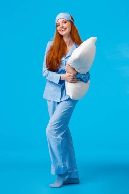 Uzun boylu, dikey stüdyo, neşeli, beyaz, kızıl, uzun saçlı, gecelikli, pijama ve uyku maskeli, yastıklı, gece yatmaya hazır, gülen, gülümseyen bir kadın.
