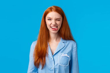 Şımarık ve heyecanlı, gece elbisesi içinde güzel saçlı bir kadın, neşeyle göz kırpan ve gülümseyen bir kamera, harika hissediyor, pozitif duygularını ifade ediyor, mavi arka planda duruyor.