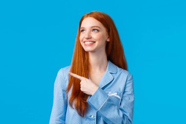 Pijama giymiş genç ve narin bir kadın, güzel bir gece elbisesi giyiyor, özel kış tatili indirimlerine bakıyor, gülümsüyor tatmin olmuş, mükemmel bir seçim yapıyor, mavi arka planda duruyor.