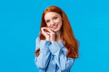 Şefkat, güzellik ve yaşam tarzı konsepti. Sevimli, mutlu, kızıl saçlı bir kadın, güzel bir gece elbisesi içinde, uyuyor, pijama giyiyor, başını eğiyor ve yüzüne dokunuyor, gülümsüyor, mavi arka plan