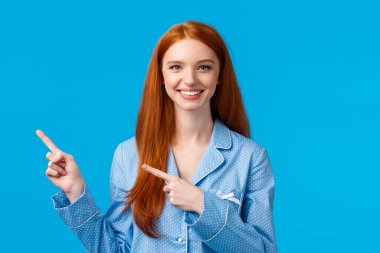 Satisfied cheerful redhead female in nightwear, pointing upper left corner, smiling friendly, help friend find place, perfect advertisement copy space, recommend product, standing blue background clipart