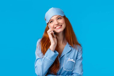 Çekici, gece boyu erkek arkadaşıyla konuşan, neşeli bir şekilde uyuyamayan, kulağının yanında akıllı telefon tutan, sırıtan, uyku maskesi takan, gece elbisesi giyen, mavi arka planda oturan bir kız.