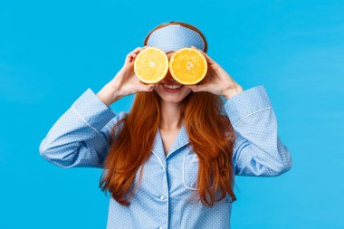 Gecelik giymiş, güzel ve tatlı kızıl saçlı kadın, uyku maskesi, iki dilim portakalı gözünün üstüne koyup gülümsüyor, sabahları maskaralık yapıyor, mavi arka planda neşe içinde dikiliyor.