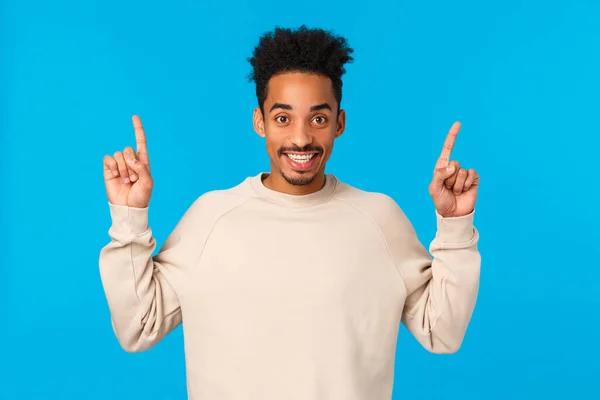 Cheerful handsome african-american young hipster guy with moustache, afro pointing fingers up, telling about promo, looking camera satisfied and beaming smile, standing blue background — Stock Photo, Image