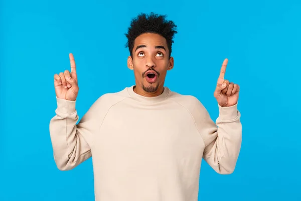 Wow awesome. Impressed good-looking african guy with moustache and afro haircut, folding lips smiling and staring, pointing up at amazing breathtaking event, advertising, blue background — Stock Photo, Image