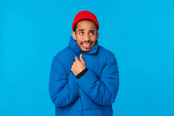 Cielos, hace frío. Joven caminando por la calle sintiendo malestar, temblando por la baja temperatura del invierno, envuélvase, cubra el cuerpo con chaqueta acolchada y gorro hipster rojo, dientes parloteando —  Fotos de Stock