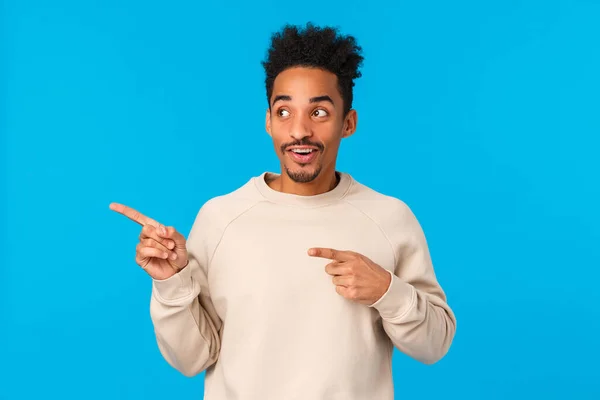 What is that. Curious and amused good-looking african american young hipster guy in white sweater, looking pointing left and smiling with pleased intrigued expression, asking about product, blue wall — Stock Photo, Image