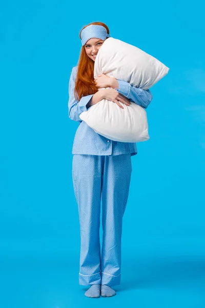 Silly romantic and cute, gentle young redhead coquettish woman in pyjama and sleep mask, hugging pillow peeking at camera with tender lovely smile, standing over blue background — Stock Photo, Image