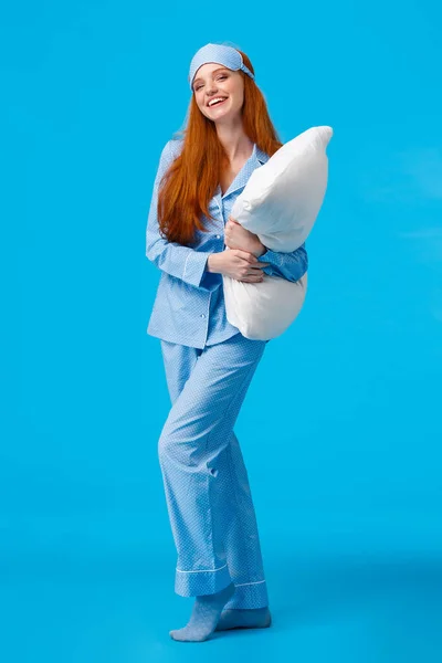 Full-length vertical studio cheerful lovely caucasian woman with ginger red long hair in nightwear, pyjama and sleep mask, hugging pillow ready sleep going bed at nigh and laughing, smiling — Stock Photo, Image