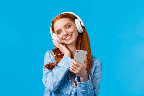 Czułość, kobiety i koncepcja stylu życia. Radosna rudowłosa kobieta w piżamie, przechylona głowa słuchająca muzyki, trzymająca smartfona i uśmiechnięta jak wzruszające słuchawki z zachwytu — Zdjęcie stockowe