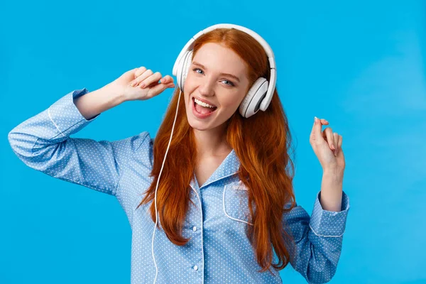 Chica despertando empezando el día con música. alegre despreocupado pelirroja adolescente bailando en ropa de dormir, levantando las manos con grandes auriculares blancos cantando a lo largo de canción favorita, lista de reproducción, fondo azul — Foto de Stock