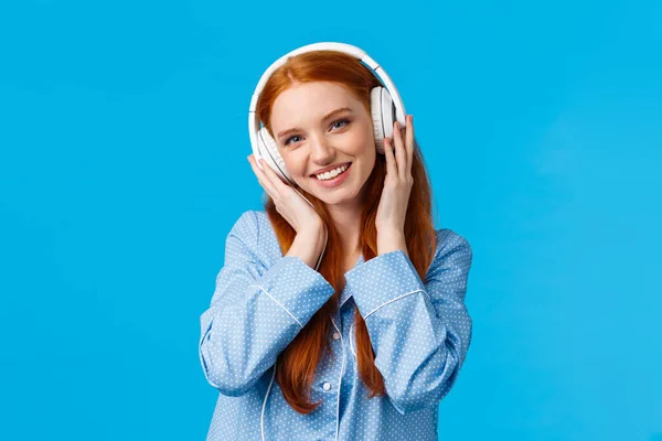 Tecnologia, estilo de vida e conceito de pessoas. Mulher ruiva bonito no pijama, ouvir música em fones de ouvido, desfrutar de boas batidas em fones de ouvido, boa qualidade, novas músicas, de pé fundo azul — Fotografia de Stock