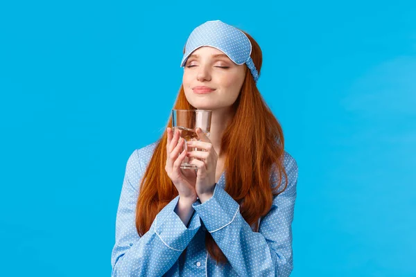 Delicious. Delighted and satisfied cute glamour redhead caucasian woman in sleep mask and nightwear drink refreshing and tasty drink, holding glass water with closed eyes and smile, blue background Royalty Free Stock Images