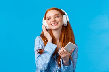 Teknoloji, müzik ve eğlence konsepti. Neşeli, çekici, kızıl saçlı, kulaklıkla müzik dinleyen, elinde akıllı bir telefon tutan, gülümseyen, mavi arka planda duran, neşeli bir kadın.
