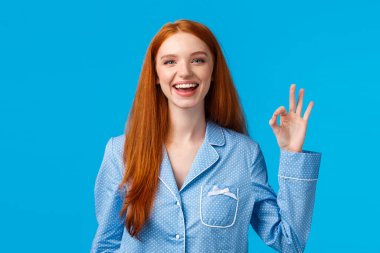 Tamam, artık endişelenmek yok. Neşeli ve rahatlamış, güler yüzlü, gece kıyafetli mutlu kızıl saçlı kız, uyumaya hazır, iyi bir jest ya da onay gösteriyor, olumlu cevap ver, mavi arka planda duruyor.