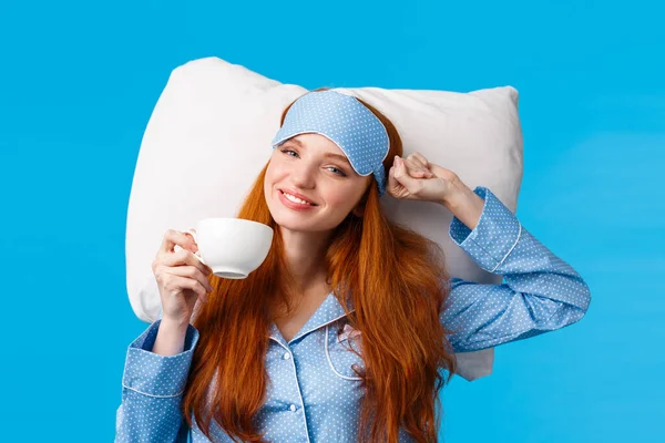 Perfect morning of princess. Cheerful, tender redhead woman with long ginger hair, wearing sleep mask and pyjama, stretching in bed feeling fresh and energized, drink morning coffee, blue background — Stock Photo, Image