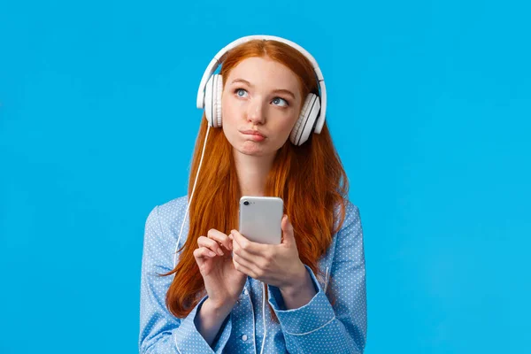 Pensativo y creativo, pensativo mujer pelirroja caucásica, chica sexy en ropa de dormir, mirando hacia arriba pensando, usando auriculares escuchar música o podcast, la celebración de teléfono inteligente, fondo azul — Foto de Stock