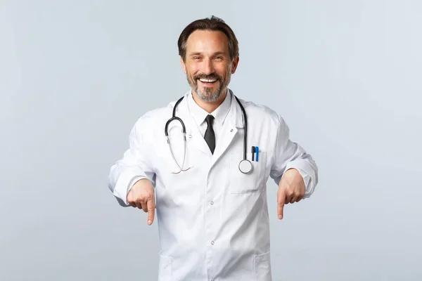 Covid-19, ξέσπασμα του ιού της στέψης, επαγγελματίες υγείας και πανδημία. Χαρούμενος χαμογελαστός άντρας γιατρός με λευκό παλτό που προσκαλεί να κάνει εξετάσεις στην κλινική, δείχνοντας τα δάχτυλα κάτω, διαφημίζοντας — Φωτογραφία Αρχείου