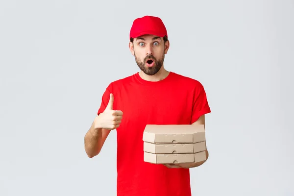 Pengiriman makanan, karantina, tinggal di rumah dan memesan konsep online. Terkejut dan takjub kurir berjenggot di t-shirt merah, topi terengah-engah terkesan, menunjukkan thumb-up, membawa pizza pesanan, latar belakang abu-abu — Stok Foto