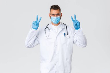 Covid-19, sağlık çalışanları, salgın hastalıklar ve virüs önleme konsepti. Beyaz takım elbiseli ve tıbbi maskeli genç iyimser doktor, lastik eldivenlerle barış işareti göster, koronavirüs salgını sırasında güçlü ol.