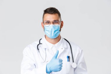 Covid-19, sağlık çalışanları, salgın hastalıklar ve virüs önleme konsepti. Tıbbi maske ve eldiven takan yakışıklı bir doktor, onay için baş parmağını kaldır, klinik hizmeti ya da ilaç tavsiye et