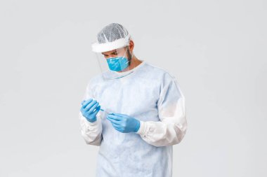 Covid-19 salgını, virüs salgını, klinik ve sağlık çalışanları konsepti. Korunma ekipmanları içinde ciddi genç bir doktor, elinde Coronavirus aşısı olan bir şırınga tutuyor, hasta hastaları tedavi ediyor.