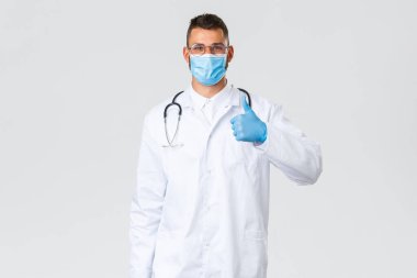 Covid-19, sağlık çalışanları, salgın hastalıklar ve virüs önleme konsepti. Hevesli, maske ve eldivenli gülümseyen doktor, steteskoplu önlükler, baş parmağını kaldırma onayı, kalite garantisi
