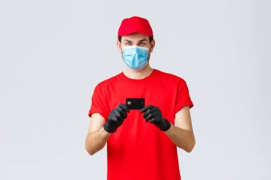 Bağlantı kurulamayan teslimat, ödeme ve kovid 19 sırasında online alışveriş, kendini karantina. Kırmızı üniforma şapkalı ve tişörtlü neşeli kurye, tıbbi maske takıyor, kredi kartını gösteren eldivenler, yemek siparişi veriyor.