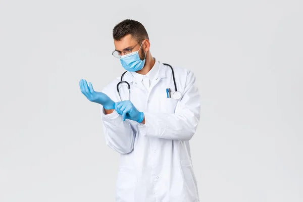 Covid-19, εργαζόμενοι στην υγειονομική περίθαλψη, πανδημία, πρόληψη της έννοιας του ιού. Νεαρός γιατρός με άσπρη ποδιά και ιατρική μάσκα που φοράει λαστιχένια γάντια για να παρατηρεί τον ασθενή στην κλινική, δουλεύοντας κατά τη διάρκεια της επιδημίας του κορωναϊού — Φωτογραφία Αρχείου