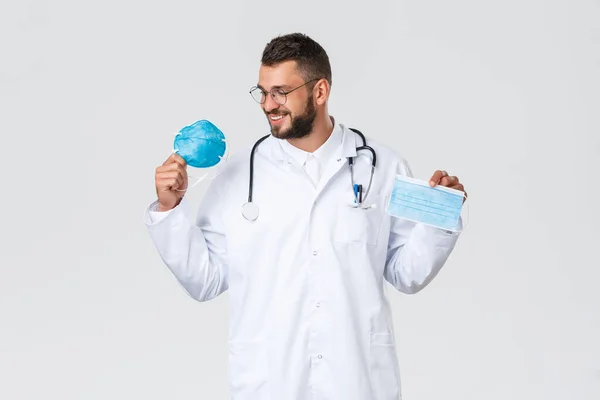 Healthcare workers, medical insurance, pandemic and covid-19 concept. Smiling handsome doctor in white coat and glasses, looking at respirator pleased, showing medical mask, grey background