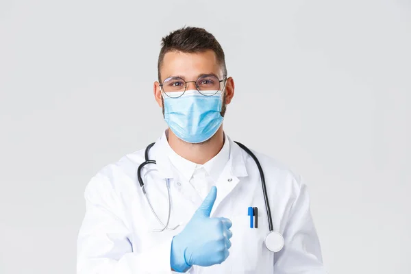 Covid-19, egészségügyi dolgozók, világjárvány és a vírus terjedésének megelőzése. Jóképű mosolygós orvos orvosi maszk és kesztyű, mutassa hüvelykujj-up jóváhagyása, ajánljuk klinika szolgáltatás vagy gyógyszer — Stock Fotó
