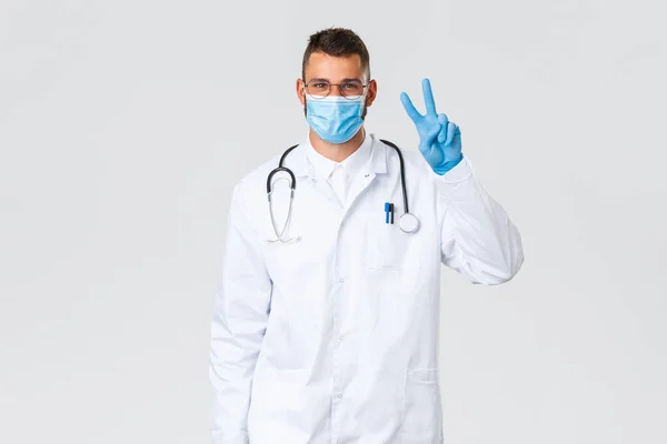 Covid-19, healthcare workers, pandemic and preventing virus concept. Friendly doctor in white scrubs, medical mask and gloves say peace, smiling keep patients safe, prevent coronavirus outbreak — Stock Photo, Image