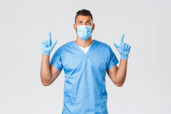Hälso- och sjukvårdspersonal, covid-19, coronavirus och förebyggande av virus koncept. Snygg seriös manlig läkare, sjuksköterska i skrubb och medicinsk mask, tittar och pekar finger upp, läsa banner promo — Stockfoto