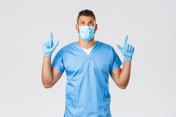 Gezondheidswerkers, covid-19, coronavirus en het voorkomen van virus concept. Knappe serieuze mannelijke arts, verpleegster in scrubs en medisch masker, kijken en wijzen vingers omhoog, het lezen van banner promo — Stockfoto