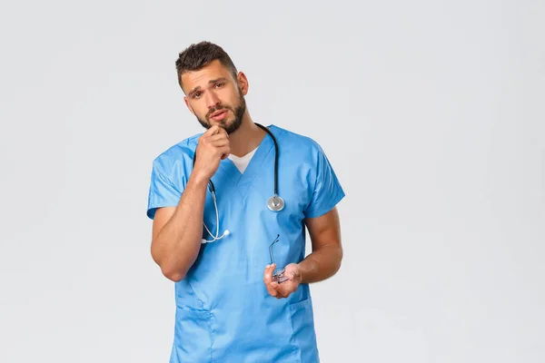 Healthcare workers, medicine, covid-19 and pandemic self-quarantine concept. Tired young doctor in blue scrubs, male nurse listenign to patient with concerned, focused face with little fatigue — Stock Photo, Image