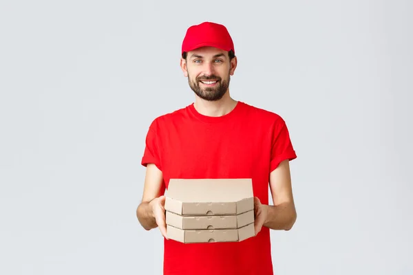 Livsmedelsleverans, karantän, stanna hemma och beställ online koncept. Le trevlig skäggig kurir i röd uniform mössa och t-shirt, ge kunder lådor med pizza, leverera ordning, grå bakgrund — Stockfoto