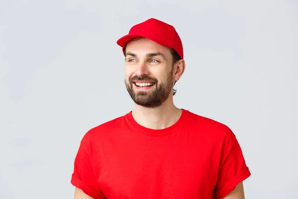 Achats en ligne, livraison en quarantaine et concept à emporter. gai barbu guy en rouge uniforme cap et t-shirt, regarder loin avec sourire heureux, lecture bannière signe, fond gris — Photo