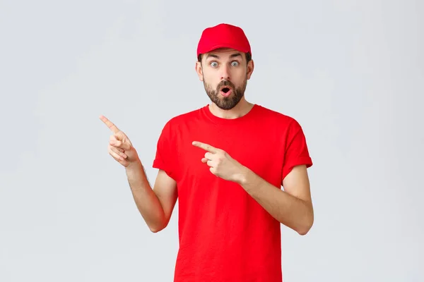 Nätshopping, leverans under karantän- och hämtningskoncept. Chockad och imponerad, kippande kurir i röd t-shirt och mössa, enhetlig service företag, pekfingrar vänster förvånad — Stockfoto