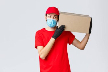 Paketler ve paket teslimatı, karantina teslimatı, nakil emirleri. Kırmızı üniformalı, eldivenli ve koruyucu maskeli bir kurye. Kutuyu müşteriye teslim et. Temassız sipariş getir.