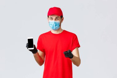 Bağlantı kurulamayan teslimat, ödeme ve kovid 19 sırasında online alışveriş, kendini karantina. Kırmızı üniformalı, eldivenli ve maskeli gülümseyen kurye, telefon ekranında sipariş bilgilerini gösteriyor, kamerayı işaret ediyor.