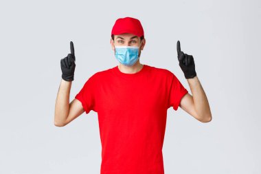 Covid-19, kendini karantina, online alışveriş ve nakliye konsepti. Mutlu teslimatçı çocuk Coronavirus tecridi sırasında kurye şirketinin reklamını yapıyor, parmaklarını kaldırıp tıbbi maske takıyor.