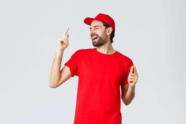 Good-looking carefree bearded man in red t-shirt and cap, uniform of employee, close eyes and dancing, pointing fingers up and singing. Delivery guy enjoying break at work, grey background — Stock Photo, Image