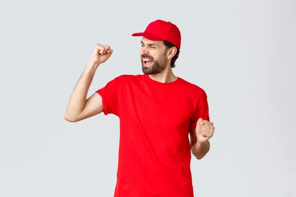 Glad och bekymmerslös ung skäggig leverans kille i röd uniform mössa och t-shirt, sjunga, skrika ja och knytnäve pump för att fira, triumfera och glädjas över goda nyheter, grå bakgrund — Stockfoto