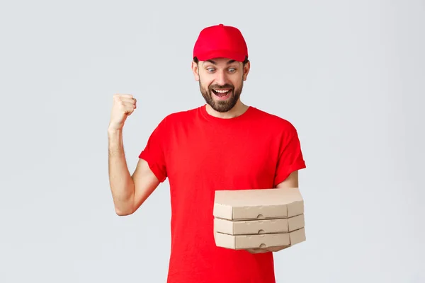 Entrega de alimentos, quarentena, ficar em casa e encomendar conceito on-line. Correios entusiasmados e felizes em camiseta e boné vermelho, regozijando-se segurando pizzas saborosas, olhe para caixas e bomba de punho, fundo cinza — Fotografia de Stock